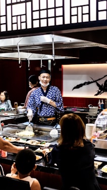 A chef wearing a blue patterned robe and white gloves is cooking in front of a seated audience at a teppanyaki restaurant. The surroundings include a variety of dishes, a grill, and Japanese-style decor with wooden panels and a large calligraphy art piece in the background. Several patrons are engaged, observing the cooking demonstration.