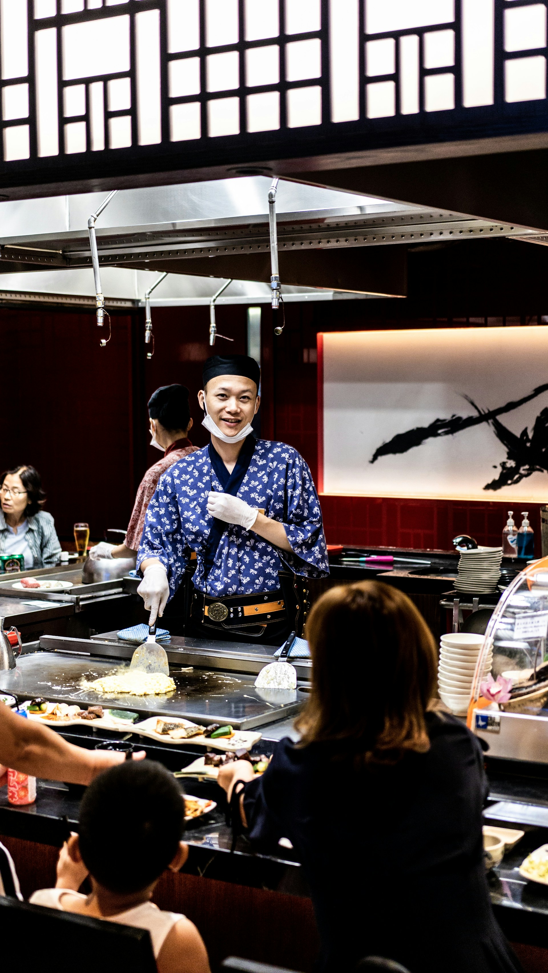 A chef wearing a blue patterned robe and white gloves is cooking in front of a seated audience at a teppanyaki restaurant. The surroundings include a variety of dishes, a grill, and Japanese-style decor with wooden panels and a large calligraphy art piece in the background. Several patrons are engaged, observing the cooking demonstration.