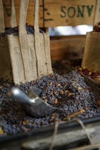 Burlap bags filled with dried lavender and herbs are displayed on a wooden stall. A metal scoop is resting amidst the lavender, suggesting a rustic market or farm setting. The earthy and aromatic atmosphere is emphasized by the presence of natural textures and colors.
