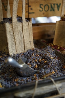 Burlap bags filled with dried lavender and herbs are displayed on a wooden stall. A metal scoop is resting amidst the lavender, suggesting a rustic market or farm setting. The earthy and aromatic atmosphere is emphasized by the presence of natural textures and colors.