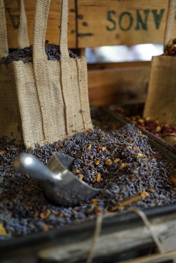 Burlap bags filled with dried lavender and herbs are displayed on a wooden stall. A metal scoop is resting amidst the lavender, suggesting a rustic market or farm setting. The earthy and aromatic atmosphere is emphasized by the presence of natural textures and colors.