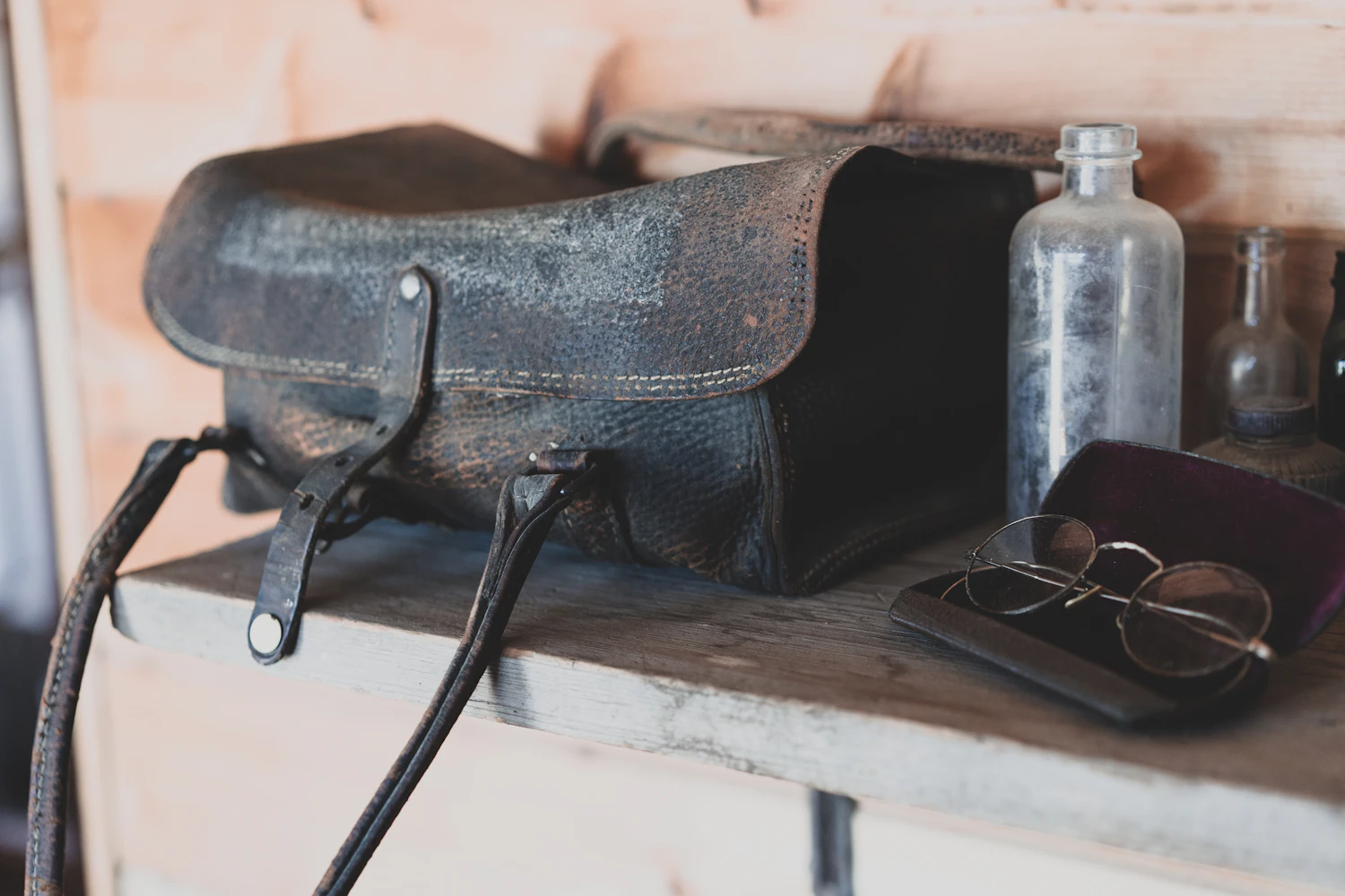 A weathered dark leather doctor's bag open on a shelf beside glass bottles and spectacles