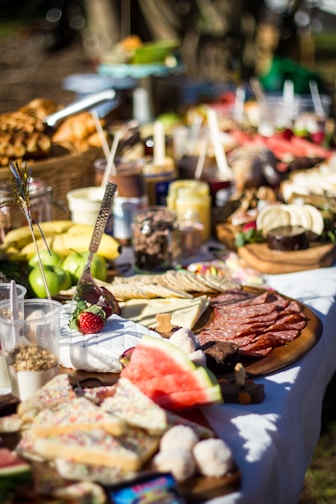 An outdoor picnic setup features a variety of foods laid out on a long table. There are sliced meats, cheeses, fresh fruits like watermelon and strawberries, and assorted bread. Beverages in clear cups and jars are visible among the spread, which is vibrant and appealing.