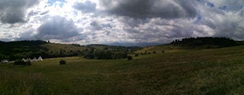 A vast, scenic landscape featuring rolling hills and a wide expanse of grassy fields under a dramatic sky filled with clouds. In the distance, several white tents are pitched among the greenery, surrounded by sparse trees and bushes. The overall scene conveys a sense of openness and tranquility.