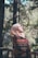 A person wearing a beige hijab and glasses is wrapped in a richly patterned shawl with red, green, and yellow colors. They appear to be outdoors, surrounded by tall trees and greenery, looking serene and contemplative with their gaze slightly upward.