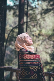 A person wearing a beige hijab and glasses is wrapped in a richly patterned shawl with red, green, and yellow colors. They appear to be outdoors, surrounded by tall trees and greenery, looking serene and contemplative with their gaze slightly upward.