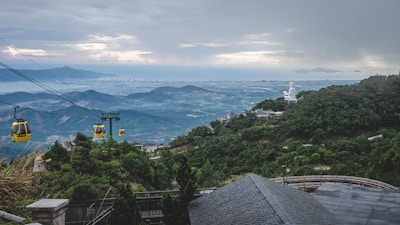 A scenic landscape featuring a panoramic view of a city by the sea, visible in the distance, with mountains in the background. A series of yellow cable cars travel through the lush green hills, and a large white statue can be seen on a hilltop.