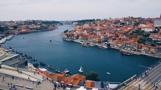 A picturesque view of a European city with a prominent river running through it. The buildings have red-tiled roofs, and small boats are floating on the water. People are visible walking along a riverside promenade, with a bridge stretching across the water in the distance.