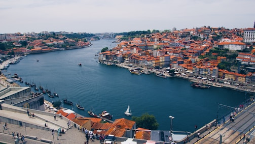 A picturesque view of a European city with a prominent river running through it. The buildings have red-tiled roofs, and small boats are floating on the water. People are visible walking along a riverside promenade, with a bridge stretching across the water in the distance.