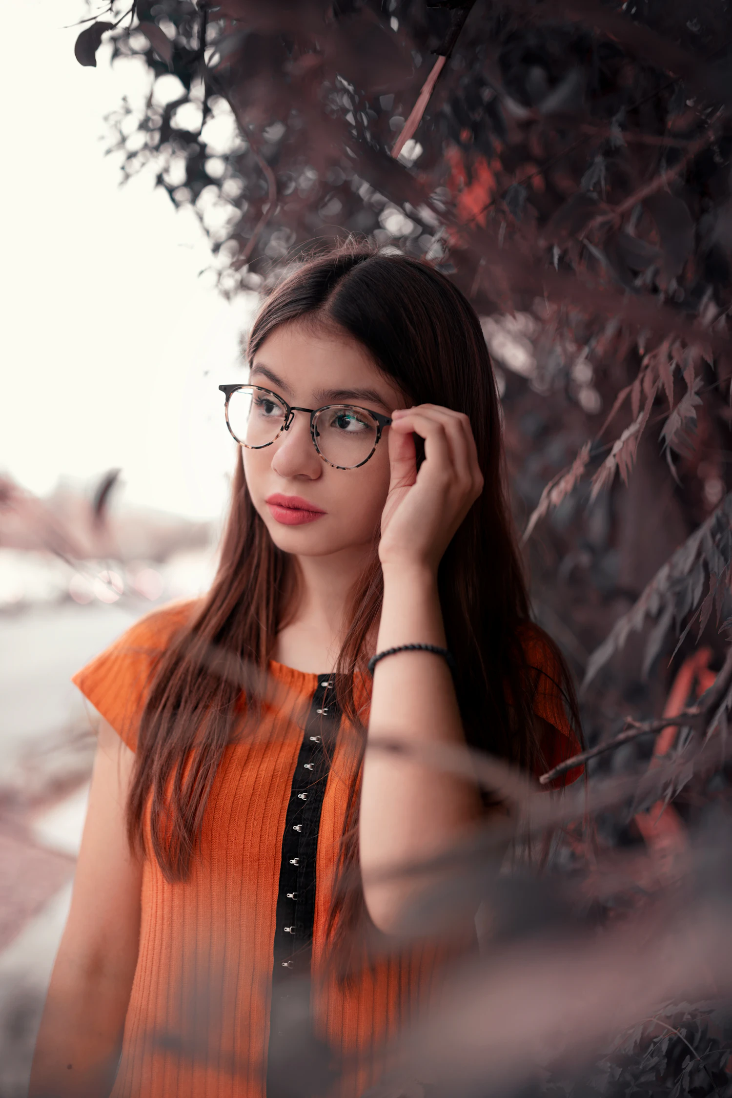 A young woman in an orange top and thin round frames pushing her hair back outdoors