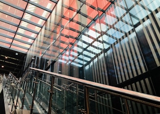 A modern architectural space featuring a staircase with glass railings, surrounded by reflective glass walls. The ceiling has a grid of alternating red and white translucent panels, creating a pattern of light and color.