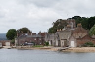 A historic waterfront estate featuring a series of stone and brick buildings with chimneys and gabled roofs. The architecture suggests an old English style, with a mix of gray and brown hues in the structures. The scene is bordered by a body of water in the foreground and lush greenery in the background, under a cloudy sky.