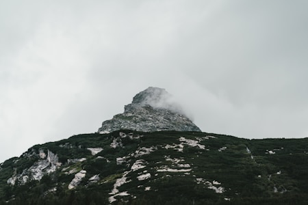 A rugged mountain peak partially shrouded in mist and clouds. The rocky outcrops and patches of greenery create an interplay of textures, giving a sense of raw natural beauty and elevation.