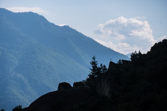 A serene landscape features a silhouette of a mountainous area with a prominent cross atop a structure on the hill. Lush greenery covers the slopes, while fluffy clouds hang in the clear sky, creating a tranquil and contemplative scene.