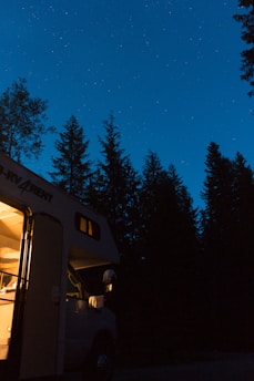 A camper van is parked among tall pine trees at night, with a warmly lit interior contrasting against the dark silhouette of the trees. The starry sky provides a serene backdrop, creating a sense of tranquility and adventure.