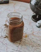 A mason jar filled with a brown beverage is placed on top of a detailed map. A small model globe and a miniature toy vehicle are nearby, indicating a theme of travel or exploration.