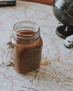 A mason jar filled with a brown beverage is placed on top of a detailed map. A small model globe and a miniature toy vehicle are nearby, indicating a theme of travel or exploration.