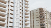 Two high-rise apartment buildings with multiple balconies are seen under an overcast sky. The structures are primarily beige with brown accents, and several balconies have satellite dishes and various items as though they are being used for storage.