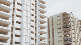 Two high-rise apartment buildings with multiple balconies are seen under an overcast sky. The structures are primarily beige with brown accents, and several balconies have satellite dishes and various items as though they are being used for storage.
