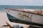 A small fishing boat is positioned on a sandy beach near the ocean. The side of the boat is painted with the phrase 'FOR SALE!' along with a phone number. The ocean is visible in the background, and the sky is partly cloudy.