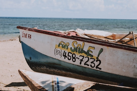 A small fishing boat is positioned on a sandy beach near the ocean. The side of the boat is painted with the phrase 'FOR SALE!' along with a phone number. The ocean is visible in the background, and the sky is partly cloudy.