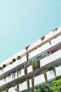 A modern building facade featuring multiple balconies adorned with lush greenery and vibrant flowers. The clean white structure contrasts with the bright green plants draping over the railings. The clear blue sky enhances the fresh and natural appearance of the scene.