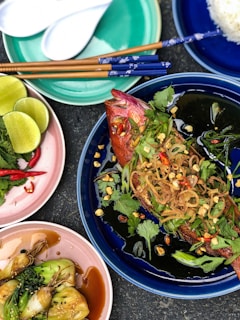A vibrant, colorful Asian meal is spread across multiple dishes. A whole cooked fish garnished with herbs, sliced chili, and crispy onions is placed on a blue plate. Additional plates hold vibrant green limes, fresh herbs, red chilies, and a side of cooked greens with sauce. Chopsticks rest on the plates, and a bowl of white rice is nearby.
