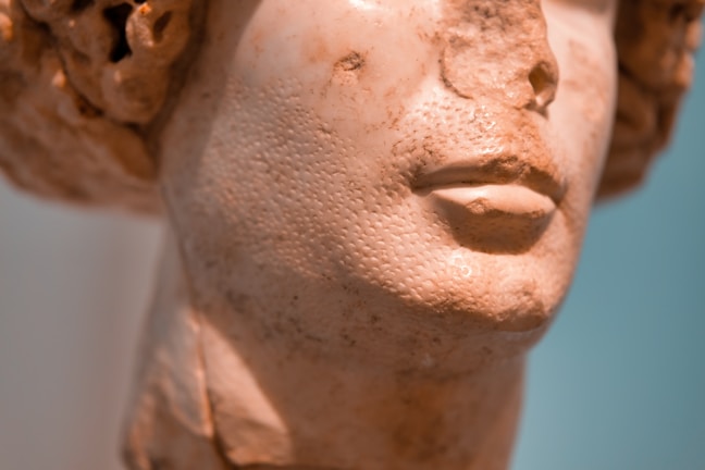 A close-up view of an old marble bust, focusing on the intricate texture and detailed craftsmanship of the sculpture's face. The surface is worn, displaying the character and age of the stone, with subtle lighting enhancing the contours and shadows of the features.