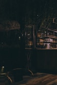 An ambient, dimly lit space with a rustic setting. The interior features glassware and metal containers on shelves, with a blend of wood and metal furnishings. The atmosphere is cozy and intimate, complemented by hanging plants and warm lighting.