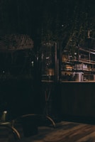 An ambient, dimly lit space with a rustic setting. The interior features glassware and metal containers on shelves, with a blend of wood and metal furnishings. The atmosphere is cozy and intimate, complemented by hanging plants and warm lighting.