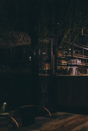 An ambient, dimly lit space with a rustic setting. The interior features glassware and metal containers on shelves, with a blend of wood and metal furnishings. The atmosphere is cozy and intimate, complemented by hanging plants and warm lighting.