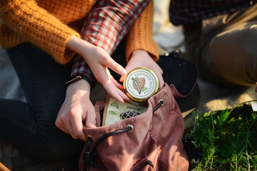 Two people are sitting outdoors with one person reaching for a jar inside a brown backpack. The jar has a lid with Cyrillic writing and an image of what appears to be pine cones. One person is wearing a watch and a textured orange sweater, while the other wears a plaid shirt. They are surrounded by greenery.