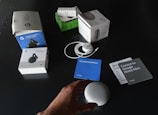 A collection of Google products and their packaging are spread out on a dark surface. Items include boxes for Google Home Mini, Chromecast, and a smart plug. Next to the boxes, an unboxed Google Home Mini, power cable, and several information cards in different colors are visible. A person's hand is reaching towards the Google Home Mini.