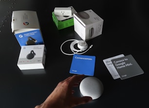 A collection of Google products and their packaging are spread out on a dark surface. Items include boxes for Google Home Mini, Chromecast, and a smart plug. Next to the boxes, an unboxed Google Home Mini, power cable, and several information cards in different colors are visible. A person's hand is reaching towards the Google Home Mini.