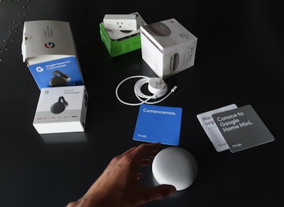 A collection of Google products and their packaging are spread out on a dark surface. Items include boxes for Google Home Mini, Chromecast, and a smart plug. Next to the boxes, an unboxed Google Home Mini, power cable, and several information cards in different colors are visible. A person's hand is reaching towards the Google Home Mini.