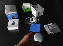 A collection of Google products and their packaging are spread out on a dark surface. Items include boxes for Google Home Mini, Chromecast, and a smart plug. Next to the boxes, an unboxed Google Home Mini, power cable, and several information cards in different colors are visible. A person's hand is reaching towards the Google Home Mini.