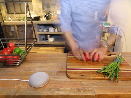 A person is chopping red bell peppers on a wooden cutting board in a kitchen. A bundle of asparagus is placed beside the board. There is a basket containing tomatoes and limes on the counter to the left, and a smart speaker is also present on the counter. The kitchen in the background has shelves with kitchenware and various items.