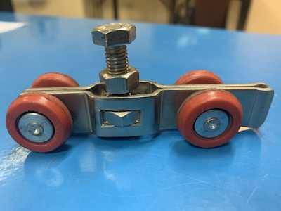 A small metal and plastic component resembling a wheeled assembly with four red wheels and a central metal structure featuring a bolt and nut. The device is placed on a light blue surface.