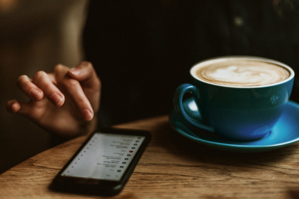 black iPhone 7 beside blue teacup with saucer and latte