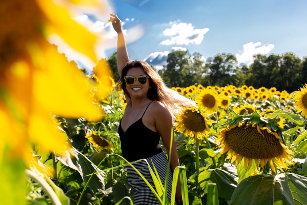 Femme en chemisier noir à bretelles spaghetti debout sur le champ de tournesol levant la main et souriant pendant la journée