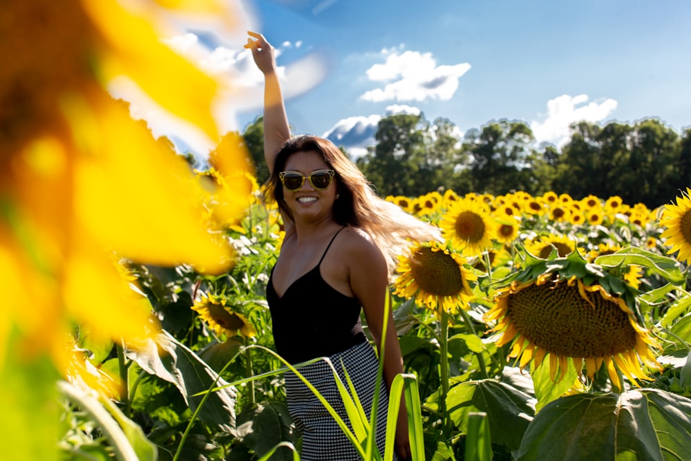 woman in black spaghetti strap blouse standing on sunflower field raising hand and smiling during daytime