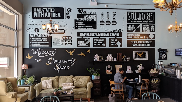 A cozy community cafe interior with soft seating including sofas and chairs, tables, and plants. The walls feature various informative and motivational posters discussing fair trade, community, and global change. A person is sitting at a table, engaged with a laptop. The decor includes chandeliers and a warm palette that creates an inviting atmosphere.