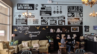 A cozy community cafe interior with soft seating including sofas and chairs, tables, and plants. The walls feature various informative and motivational posters discussing fair trade, community, and global change. A person is sitting at a table, engaged with a laptop. The decor includes chandeliers and a warm palette that creates an inviting atmosphere.