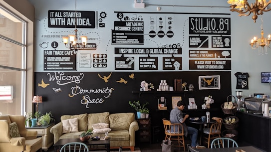 A cozy community cafe interior with soft seating including sofas and chairs, tables, and plants. The walls feature various informative and motivational posters discussing fair trade, community, and global change. A person is sitting at a table, engaged with a laptop. The decor includes chandeliers and a warm palette that creates an inviting atmosphere.