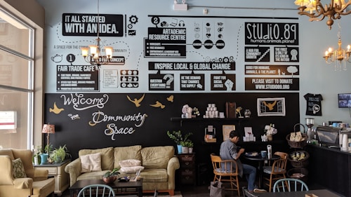 A cozy community cafe interior with soft seating including sofas and chairs, tables, and plants. The walls feature various informative and motivational posters discussing fair trade, community, and global change. A person is sitting at a table, engaged with a laptop. The decor includes chandeliers and a warm palette that creates an inviting atmosphere.