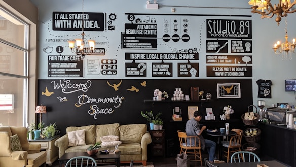 A cozy community cafe interior with soft seating including sofas and chairs, tables, and plants. The walls feature various informative and motivational posters discussing fair trade, community, and global change. A person is sitting at a table, engaged with a laptop. The decor includes chandeliers and a warm palette that creates an inviting atmosphere.