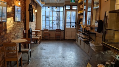 A cozy café with rustic brick walls and wooden furniture. The room contains small round tables with chairs along the sides. Large windows at the far end allow natural light to fill the space, showcasing shelves stacked with cups and glass jars. Neutral and warm lighting creates an inviting atmosphere.