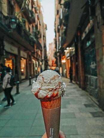 A waffle cone topped with creamy white ice cream drizzled with red syrup is being held in a narrow, cobblestone street lined with tall buildings and greenery.