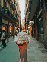 A waffle cone topped with creamy white ice cream drizzled with red syrup is being held in a narrow, cobblestone street lined with tall buildings and greenery.