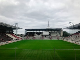 An empty football stadium with green grass on the field and stands surrounding it on three sides. There is a large banner with the message 'Kein Mensch ist illegal', accompanied by a black heart in the central stand. The architecture is modern with partial roofing over the stands, and the sky is overcast.